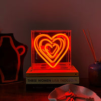 A glowing red heart-shaped neon light inside a clear acrylic box, displayed on a table with decorative items around it.